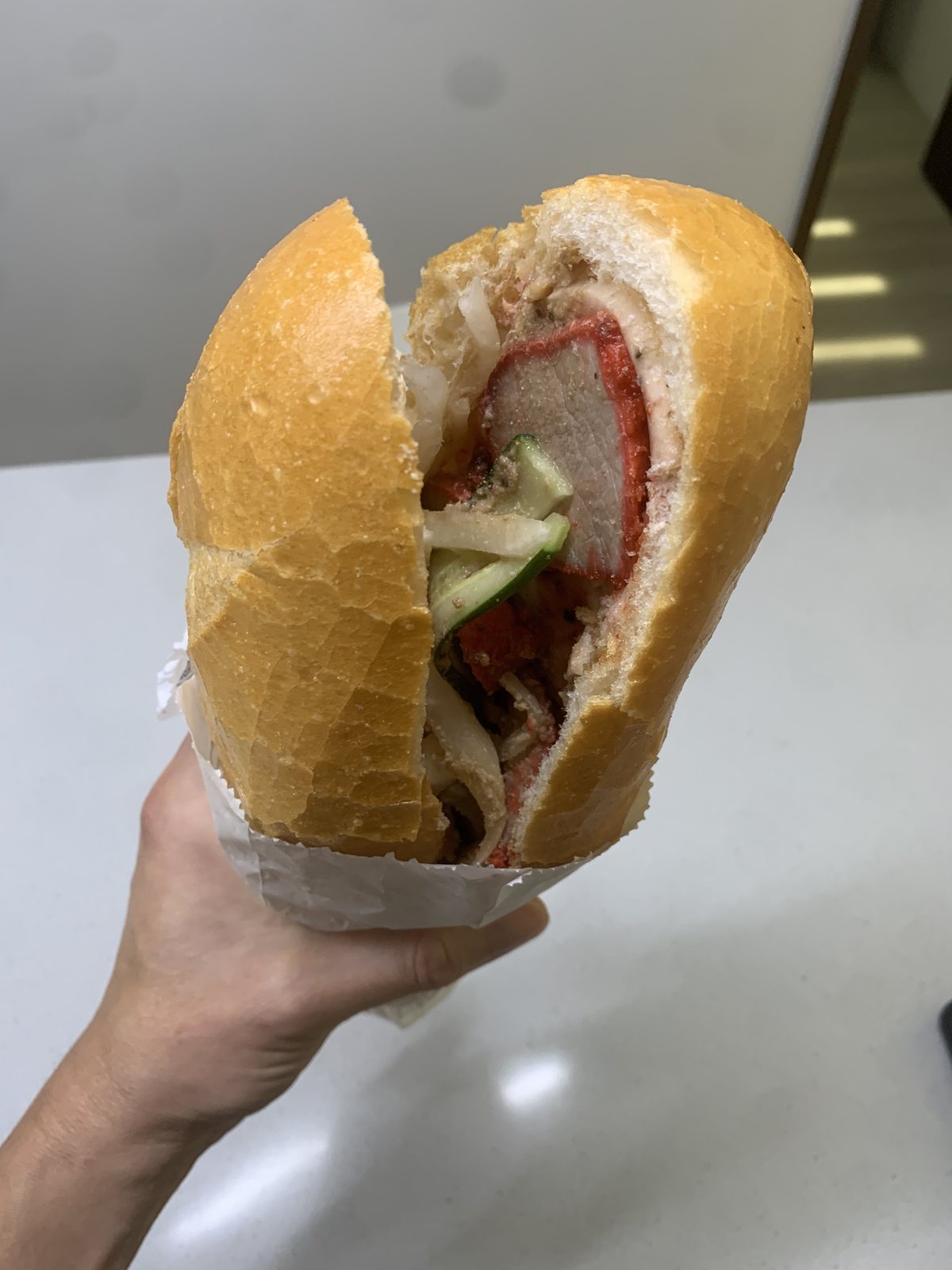 A hand holding a char siu pork banh mi cut open in white paper, with the baguette, pork and cucumber visible.